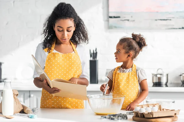 Mãe afro-americana lendo livro de receitas e filha batendo ovos para massa na cozinha — Fotografia de Stock