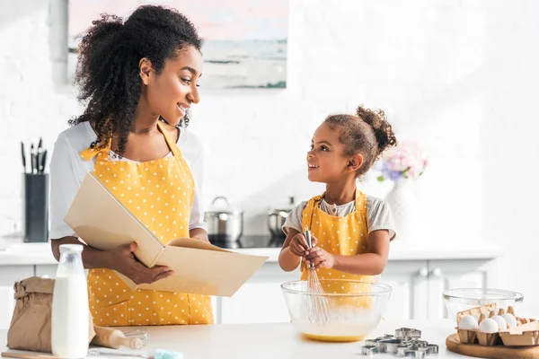 Afro-americana madre in possesso di ricettario e figlia preparare pasta in cucina, guardandosi — Foto stock