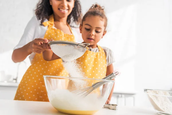 Cropped image of african american mother and daughter preparing dough and sieving flour in kitchen — Stock Photo