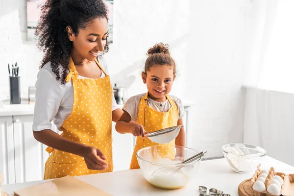 Happy african american mother and daughter preparing dough and sieving flour together in kitchen — Stock Photo
