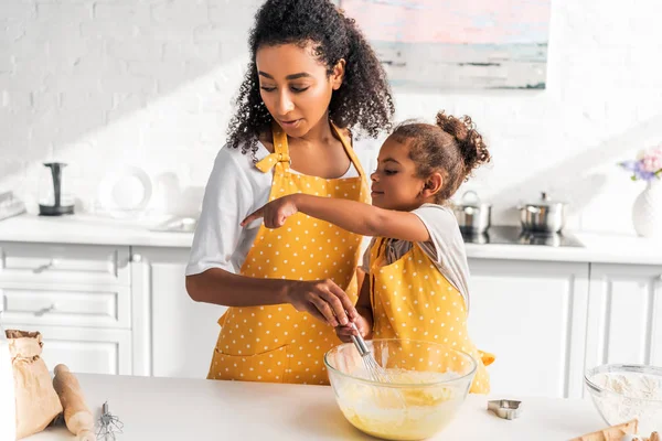 Afro-americana filha preparando e batendo massa, apontando em algo para a mãe na cozinha — Fotografia de Stock