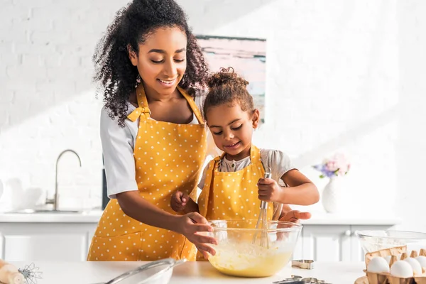 Smiling african american mother helping daughter preparing and whisking dough in kitchen — Stock Photo