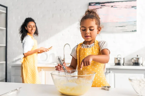 Afro-americana filha preparando e batendo massa, colocando o dedo em tigela na cozinha — Fotografia de Stock