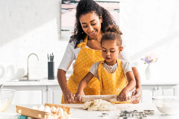 Afro-américaine mère aider fille rouler la pâte avec rouleau à pâtisserie dans la cuisine — Photo de stock