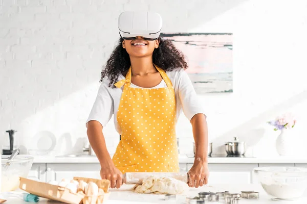 Smiling african american girl in apron and virtual reality headset rolling dough in kitchen — Stock Photo