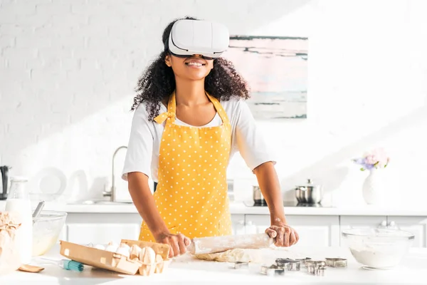 African american girl in apron and virtual reality headset rolling dough with rolling pin in kitchen — Stock Photo