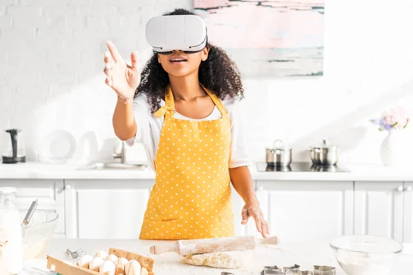 Excited african american girl in apron and virtual reality headset reaching hand at table with dough in kitchen — Stock Photo