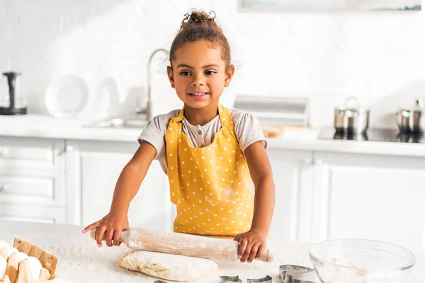 Sorridente adorabile africano americano bambino rotolamento pasta con mattarello in cucina — Foto stock