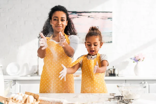 Madre e hija afroamericana preparando masa y divirtiéndose con harina en la cocina - foto de stock