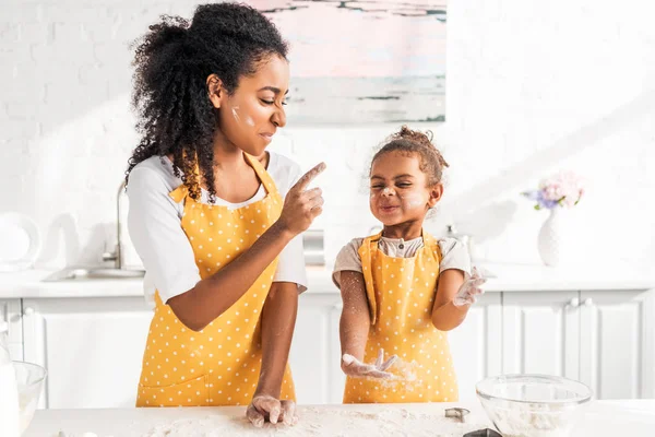Grimacing african american mother and daughter preparing dough and having fun with flour in kitchen — Stock Photo