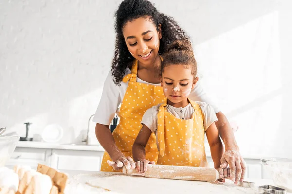 Sorrindo afro-americana mãe ajudando filha rolando massa com rolo de pino na cozinha — Fotografia de Stock