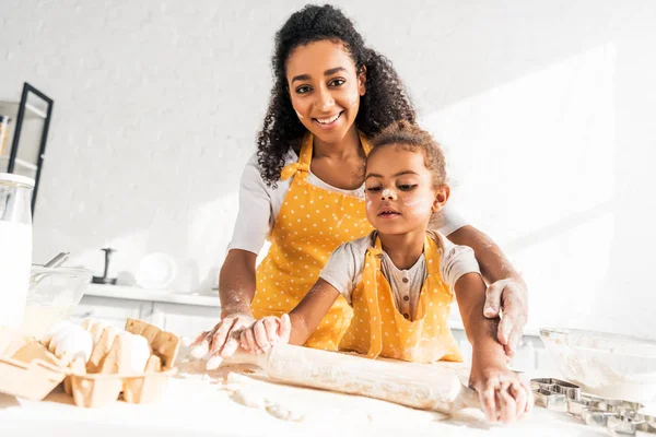 Feliz africano americano madre ayudar hija balanceo masa con rodillo en cocina - foto de stock