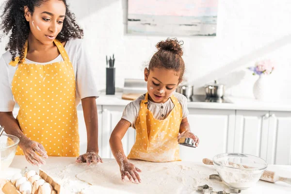 Madre e hija afroamericana en delantales preparando galletas con moldes en la cocina - foto de stock