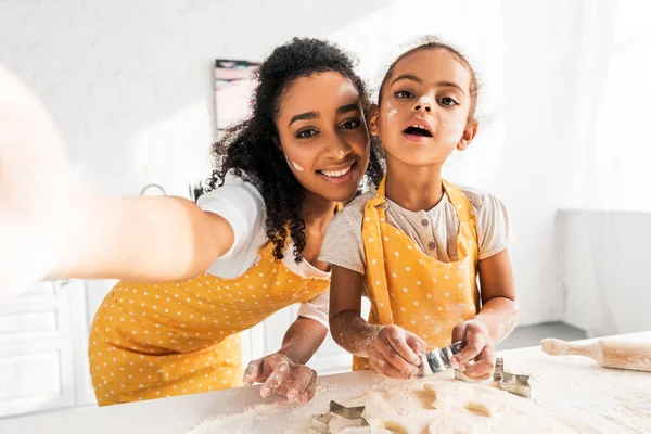 Point de vue de la caméra de sourire afro-américaine mère et fille dans des tabliers préparant des biscuits dans la cuisine — Photo de stock