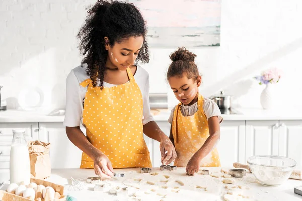 Madre e hija afroamericana en delantales amarillos preparando galletas con moldes en la cocina - foto de stock