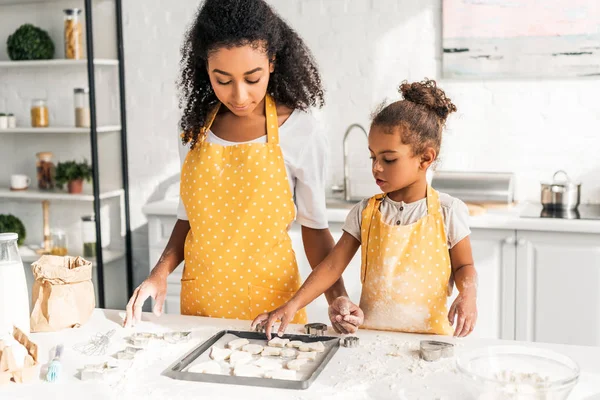 Afro-americana mãe e filha em aventais amarelos olhando para cookies na bandeja na cozinha — Fotografia de Stock