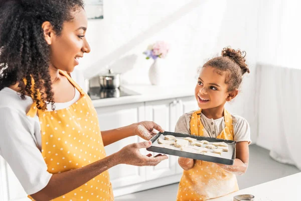 African american mother and daughter holding tray with unbaked cookies in kitchen and looking at each other — Stock Photo