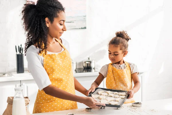 Madre e hija afroamericana en delantales sosteniendo bandeja con galletas sin cocer en la cocina — Stock Photo