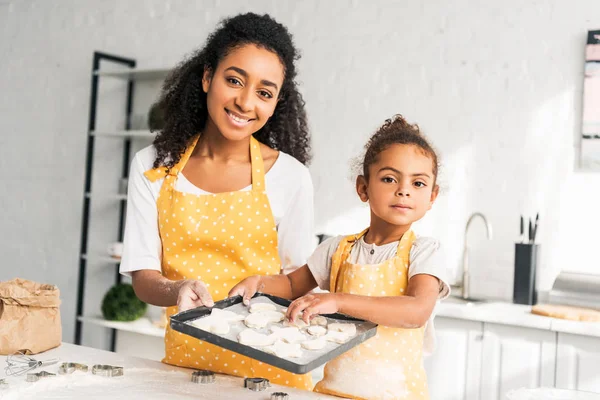 Sonriente afroamericana madre e hija sosteniendo bandeja con galletas sin cocer en la cocina y mirando a la cámara - foto de stock