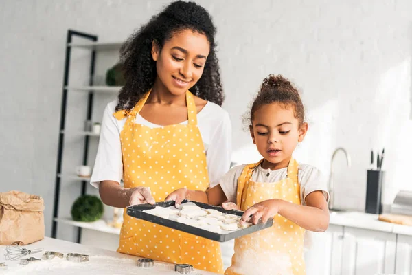 Alegre africana americana madre e hija sosteniendo bandeja con galletas sin cocer en la cocina - foto de stock