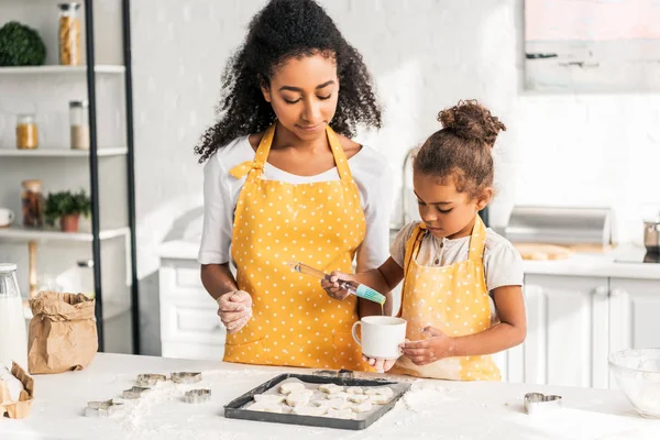 Afro-américaine mère et fille appliquant de l'huile sur les biscuits non cuits dans la cuisine — Photo de stock