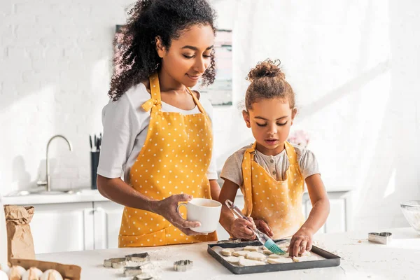 Madre e hija afroamericana en delantales amarillos aplicando aceite en galletas sin cocer en la cocina - foto de stock