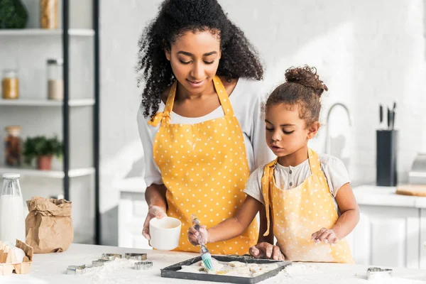 Atractiva madre afroamericana y adorable hija aplicando aceite en galletas sin cocer en la cocina - foto de stock
