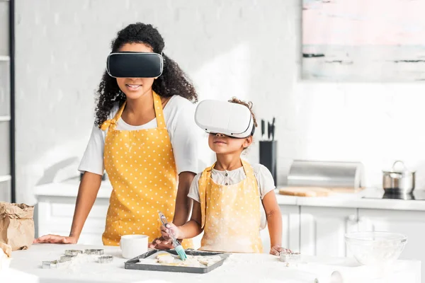 African american mother and daughter applying oil on unbaked cookies with virtual reality headsets in kitchen — Stock Photo