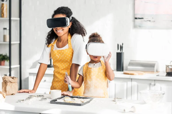 Afro-américaine mère et fille dans des tabliers appliquant de l'huile sur les biscuits non cuits avec des casques de réalité virtuelle dans la cuisine — Photo de stock