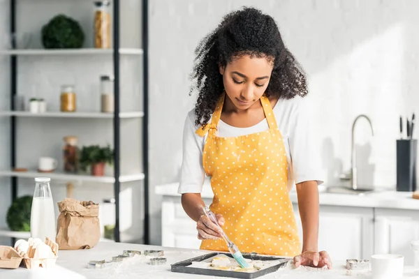 Attraktive afrikanisch-amerikanische Mädchen in Schürze Anwendung Öl auf ungebackene Kekse in der Küche — Stockfoto