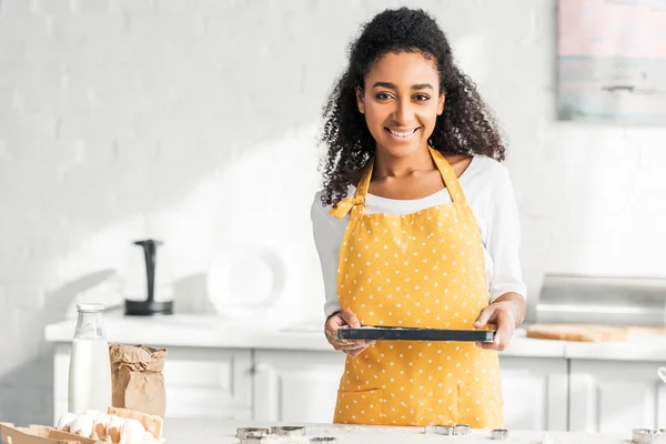 Sonriente atractiva afroamericana chica en delantal celebración bandeja con galletas sin cocer en la cocina - foto de stock