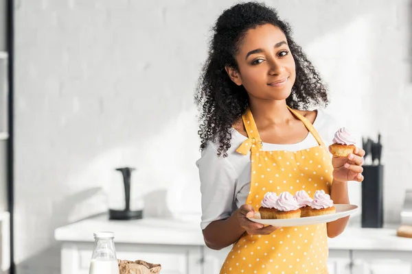 Alegre bela menina americana africana em avental segurando cupcakes caseiros e olhando para a câmera na cozinha — Fotografia de Stock