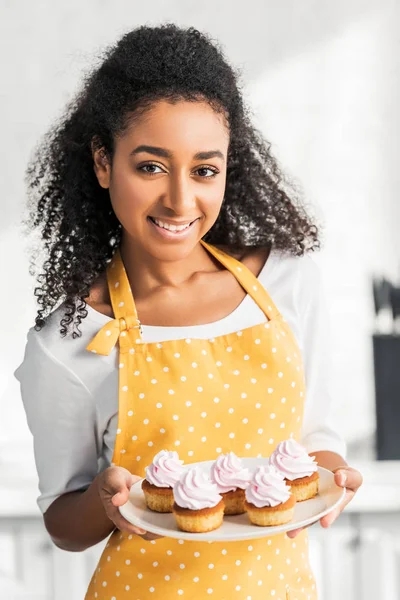 Portrait of smiling attractive african american girl in apron holding homemade cupcakes and looking at camera in kitchen — Stock Photo