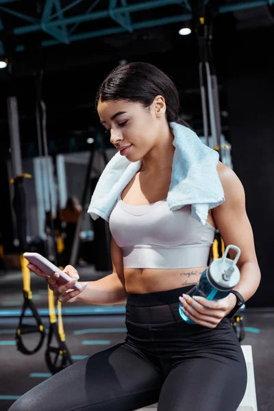 Hermosa chica deportiva usando dispositivo digital mientras sostiene la botella de deporte en el gimnasio - foto de stock