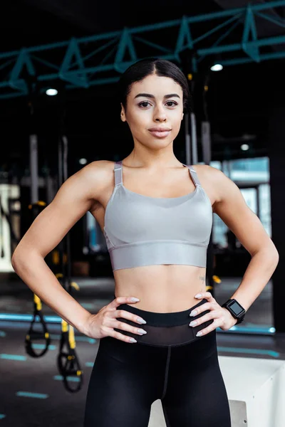 Attractive sportive girl standing akimbo while looking at camera in fitness gym — Stock Photo