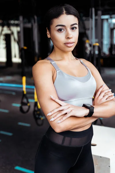 Menina esportiva bonita com braços cruzados olhando para a câmera enquanto em pé no ginásio de fitness — Fotografia de Stock