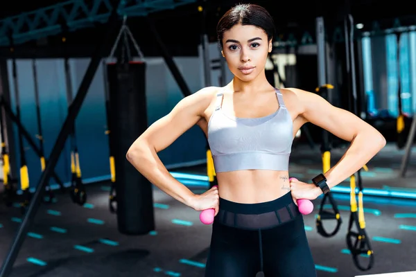 Sportive girl standing akimbo with dumbbells in fitness gym — Stock Photo