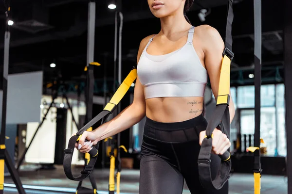 Vue recadrée d'une jolie fille sportive faisant de l'exercice avec des bretelles de suspension dans une salle de sport — Photo de stock