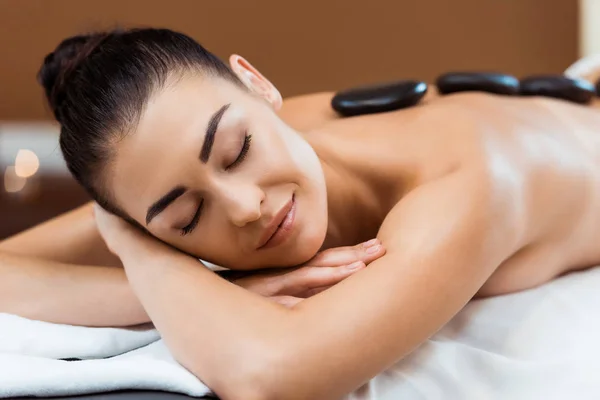 Jeune femme souriante aux yeux fermés profitant d'un massage aux pierres chaudes dans un salon de spa — Photo de stock
