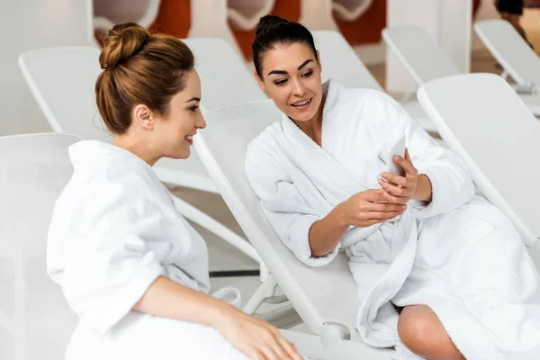 Happy young women looking at smartphone while lying together on sunbeds in spa — Stock Photo