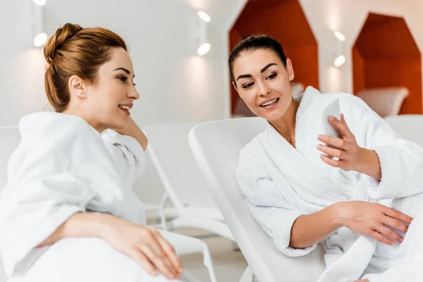 Mujeres jóvenes sonrientes mirando el teléfono inteligente mientras descansan juntas en tumbonas en el spa - foto de stock