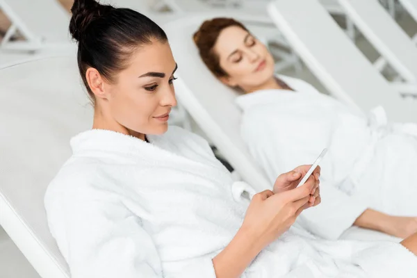 Mujer joven sonriente en albornoz usando teléfono inteligente mientras descansa en la cama de sol en el spa - foto de stock