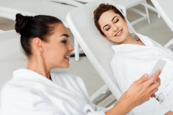 Mujeres jóvenes sonrientes en batas de baño con teléfono inteligente y hablar mientras se relaja en el spa - foto de stock