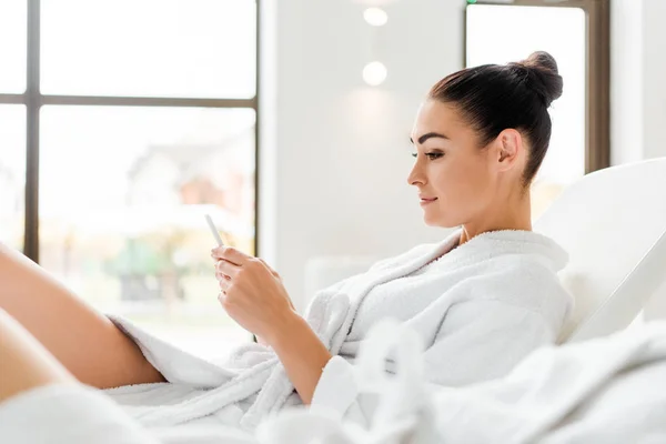 Side view of smiling young woman in bathrobe using smartphone while resting in spa — Stock Photo