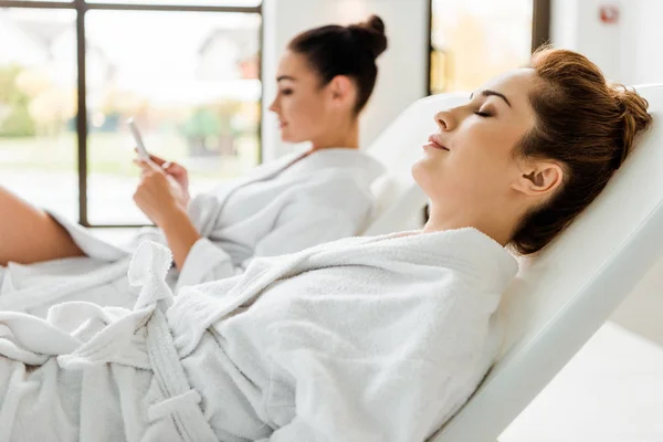 Vue latérale de jeunes femmes en peignoirs se détendre sur des chaises longues dans le spa — Photo de stock