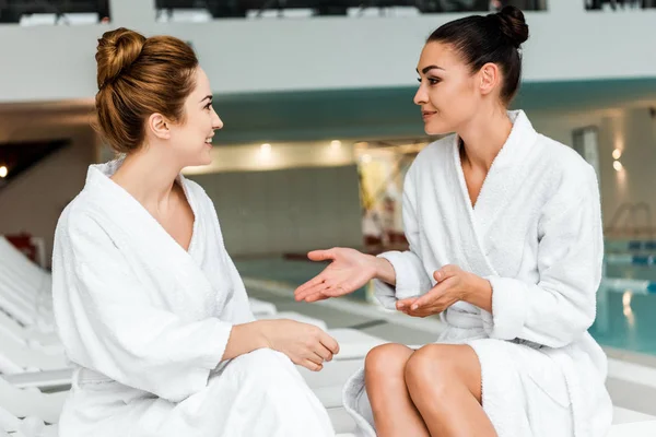 Sonrientes mujeres jóvenes en albornoces hablando mientras se relajan juntas en el spa - foto de stock