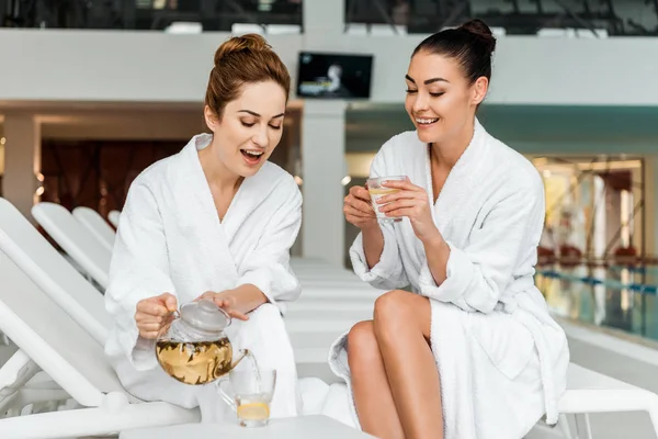Mujeres jóvenes felices en batas de baño beber té de hierbas y hablar en el spa - foto de stock