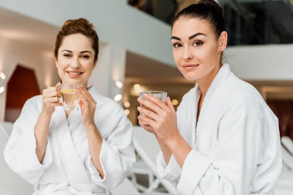 Hermosas mujeres jóvenes en albornoces sosteniendo tazas con té de hierbas y sonriendo a la cámara en el spa - foto de stock