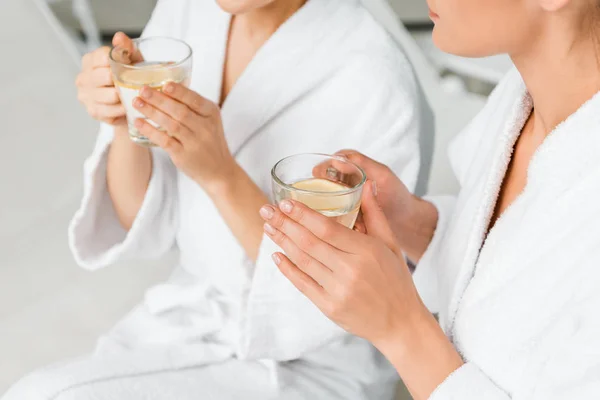 Foto recortada de mujeres jóvenes en albornoces sosteniendo tazas con té de hierbas y limón mientras descansa en el spa - foto de stock