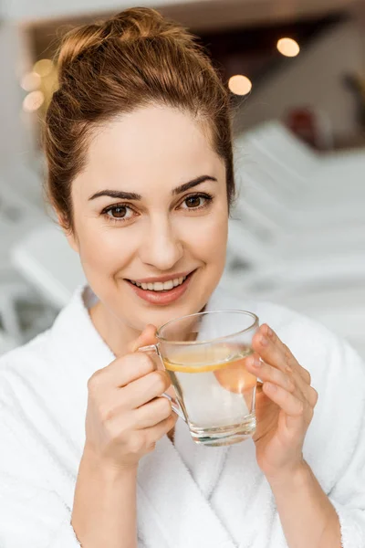 Felice giovane donna in possesso di tazza con bevanda a base di erbe e sorridente alla fotocamera in spa — Foto stock